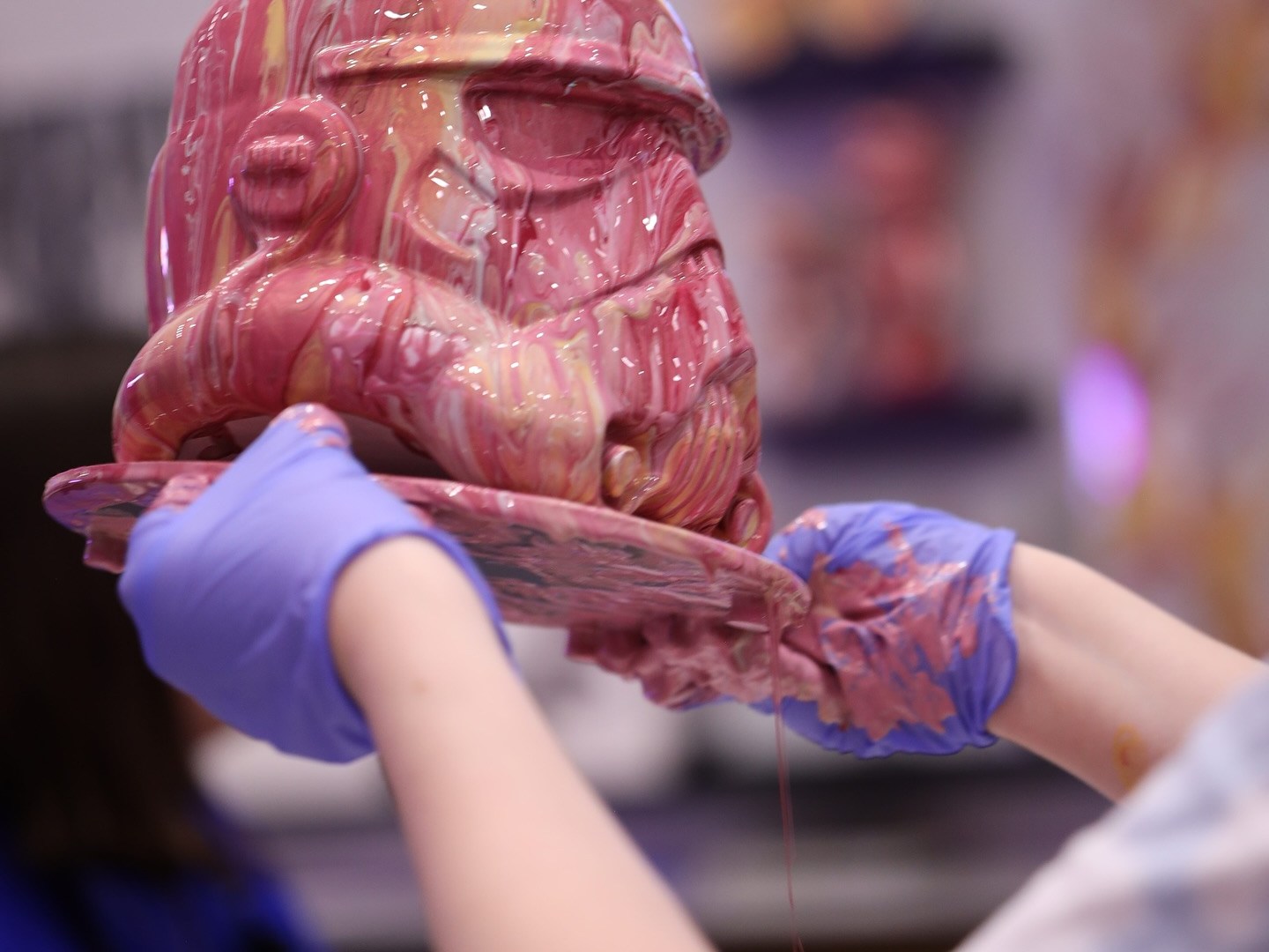 Person holding a pink marbled helmet with gloved hands in a blurred background.