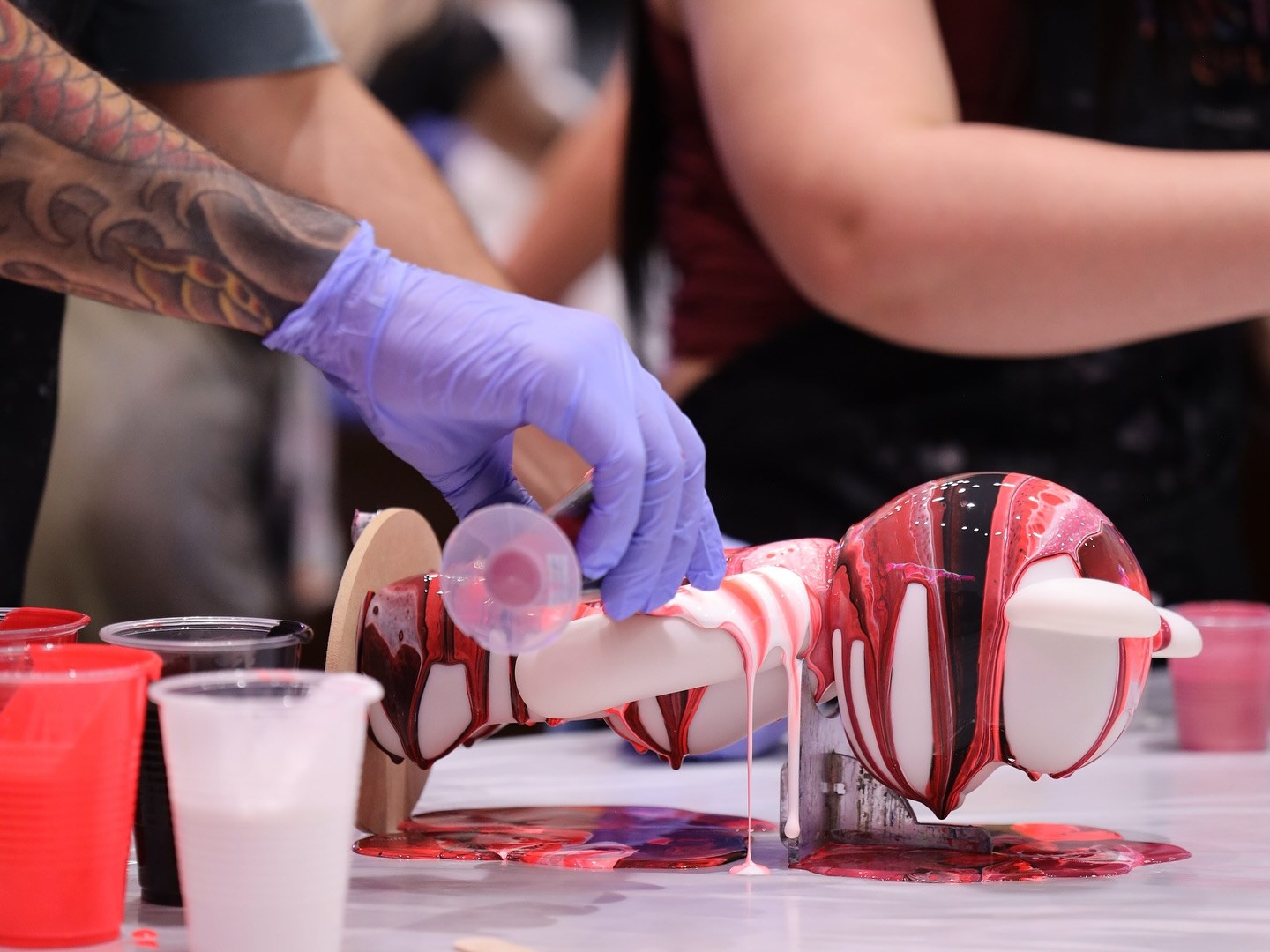 Person pours paint over a sculpture in a colorful art activity, with cups and gloves visible.