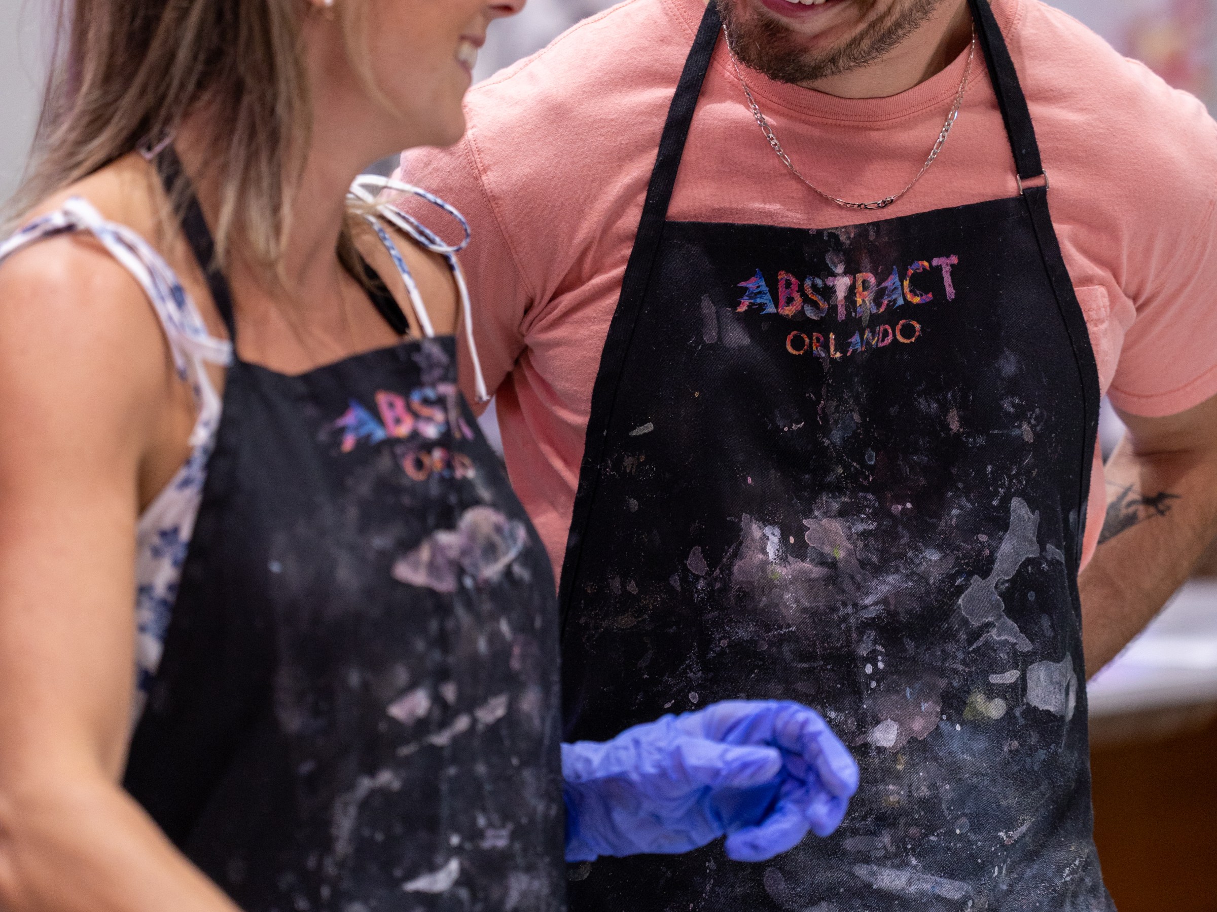 Two people in aprons, smiling at each other in an art studio with paint supplies.