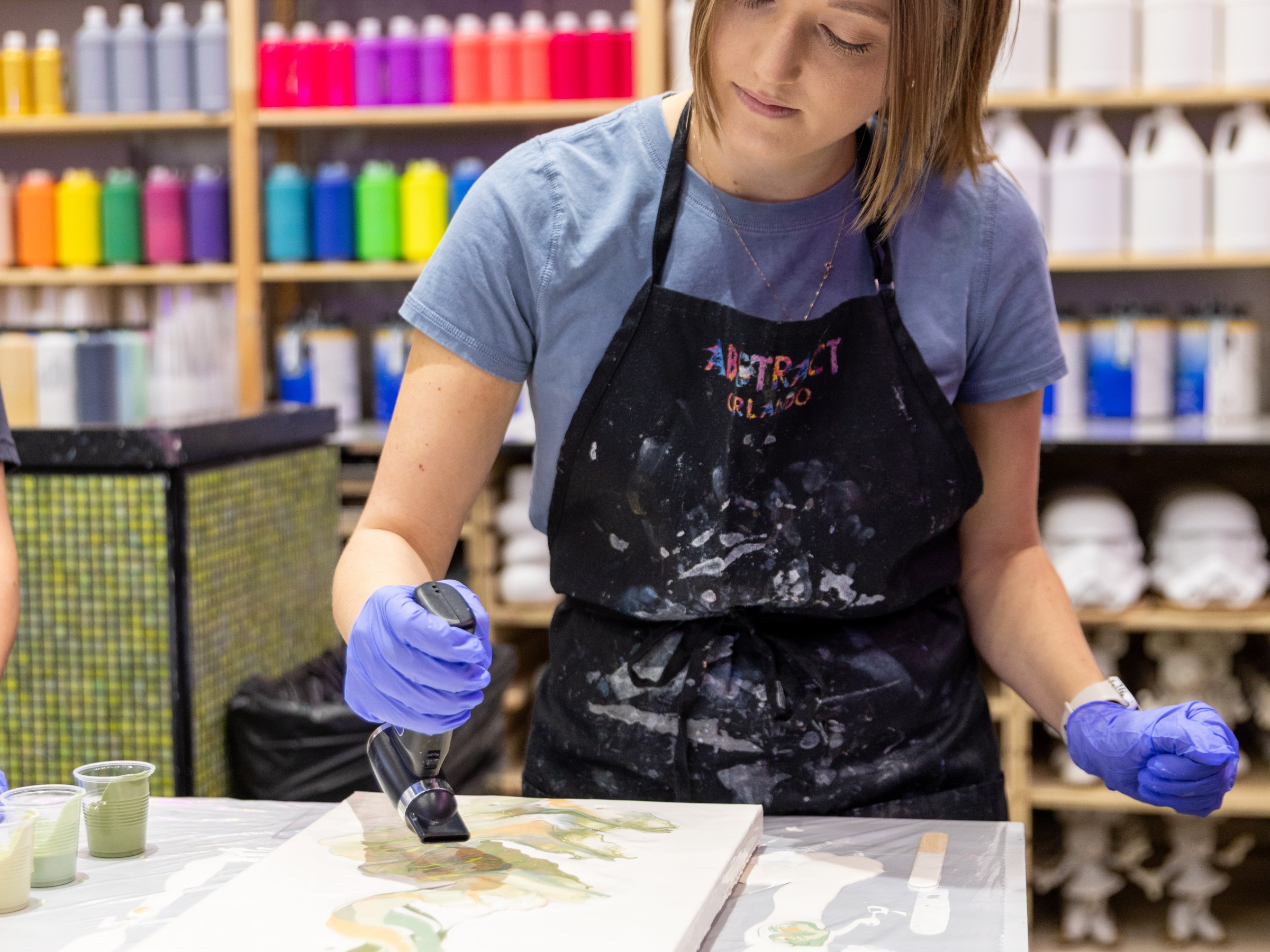 Person painting with heat gun in art store, shelves of colorful bottles behind.