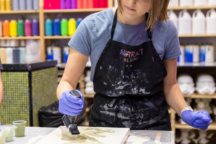 Person painting with heat gun in art store, shelves of colorful bottles behind.