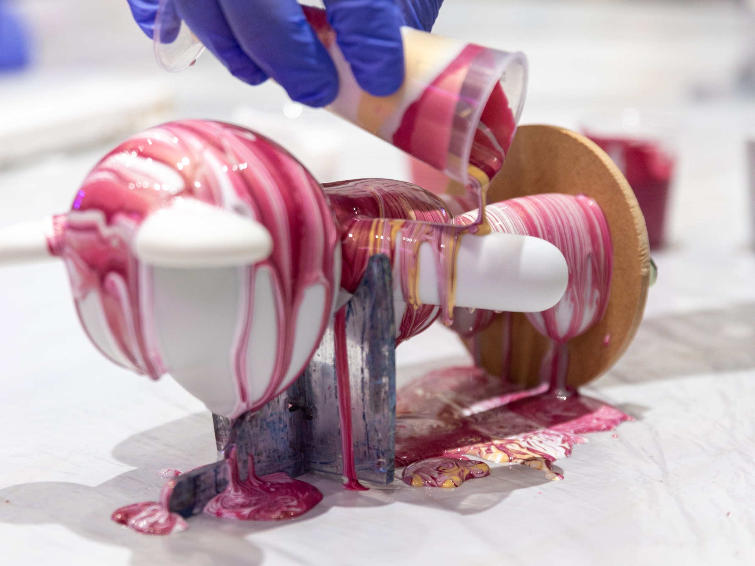 Gloved hands pour pink paint over a spinning object, creating a marbled effect.