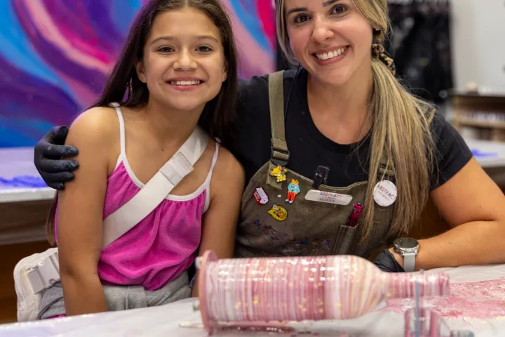 Two smiling people posing in front of colorful abstract artwork with a paint-covered bottle on a table.