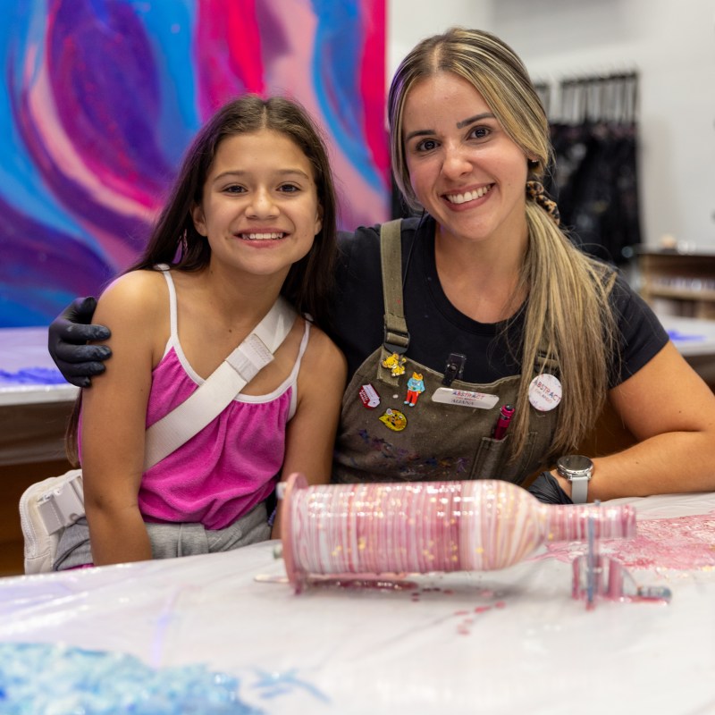Two smiling people posing in front of colorful abstract artwork with a paint-covered bottle on a table.