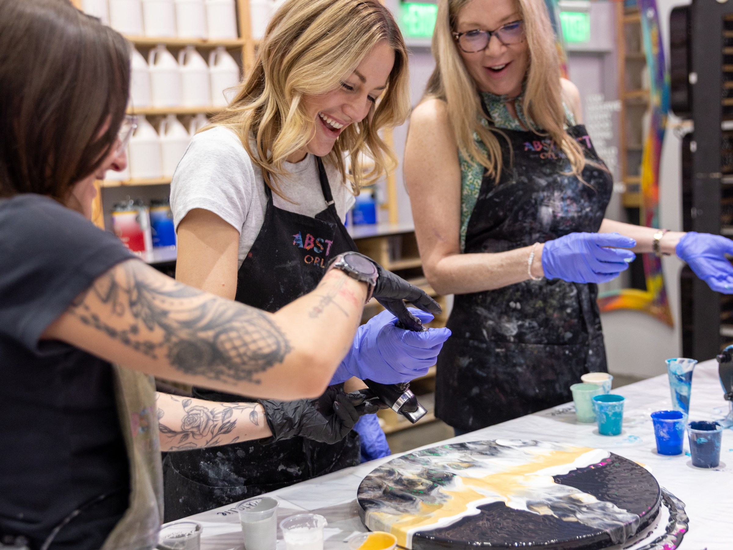 Three women in aprons painting with vibrant colors at art studio.