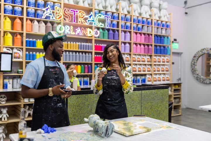 People laughing at an art studio with colorful paint bottles and 'ABSTRACT ORLANDO' sign in the background.