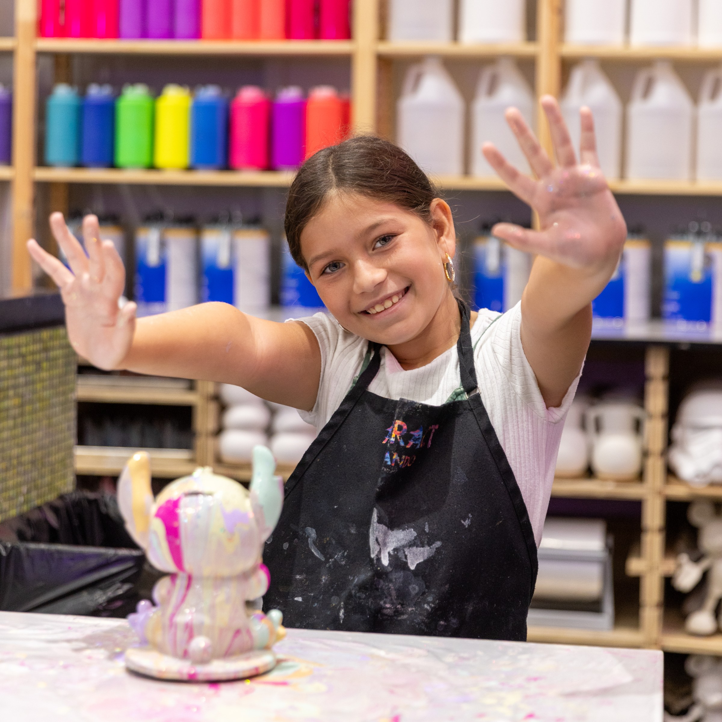 Smiling child in art apron shows painted hands, colorful shelves in background.