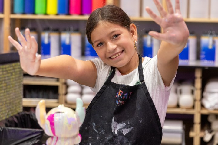 Smiling child in art apron shows painted hands, colorful shelves in background.