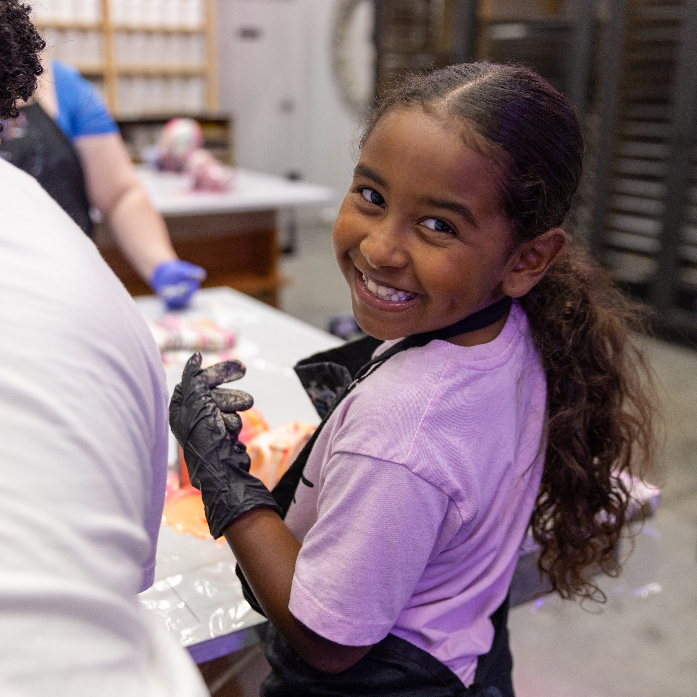 Smiling child with curly hair wearing black gloves and apron, standing by a table with colorful jars.
