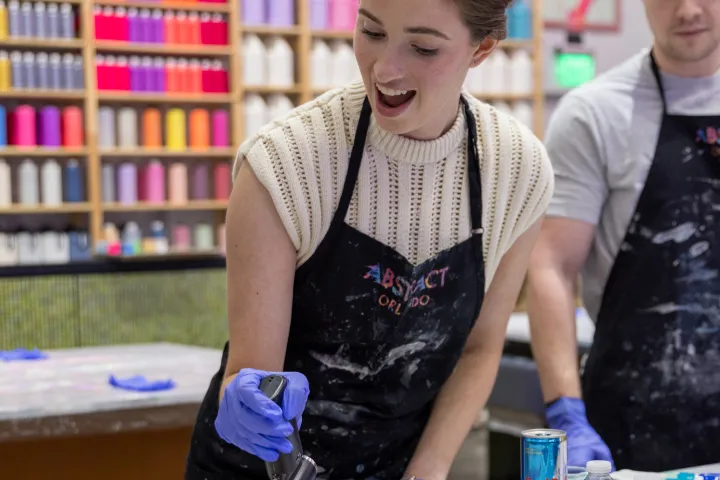 Person wearing gloves using a tool to create art on a canvas in a colorful studio.