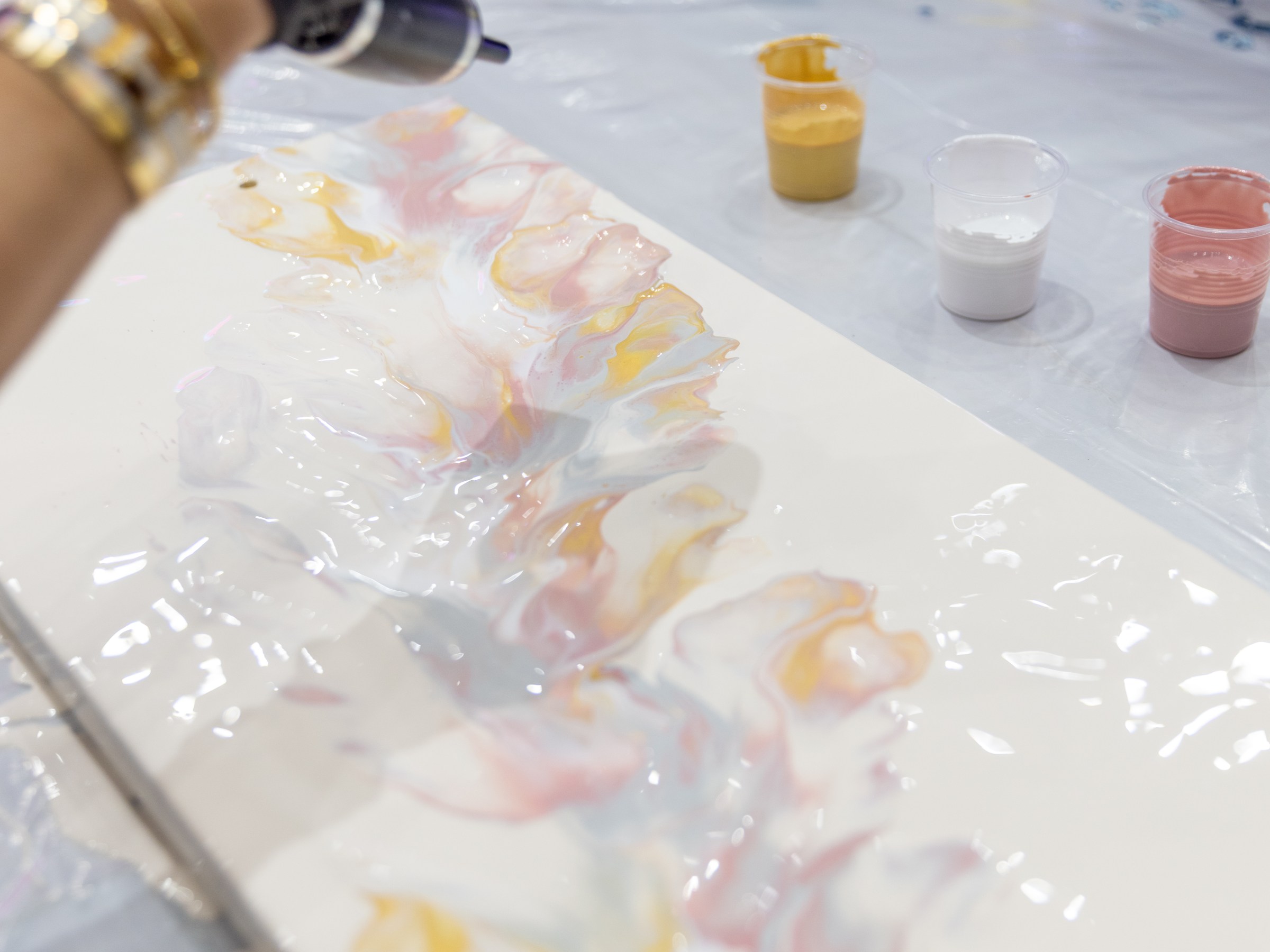 Person using a heat gun on colorful resin art with paint cups nearby.