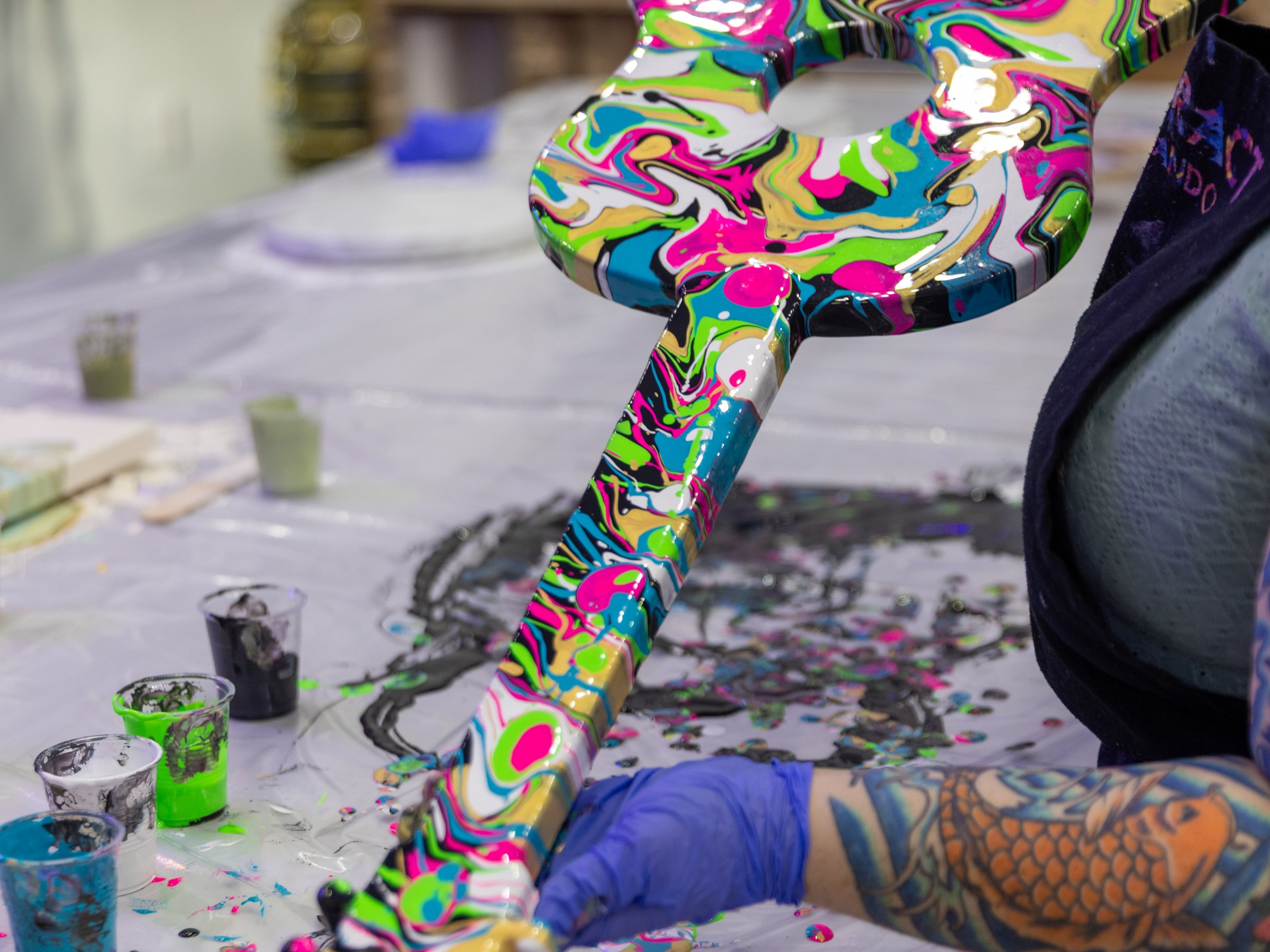 Person paints guitar body with colorful, abstract swirls on a table with paint cups.