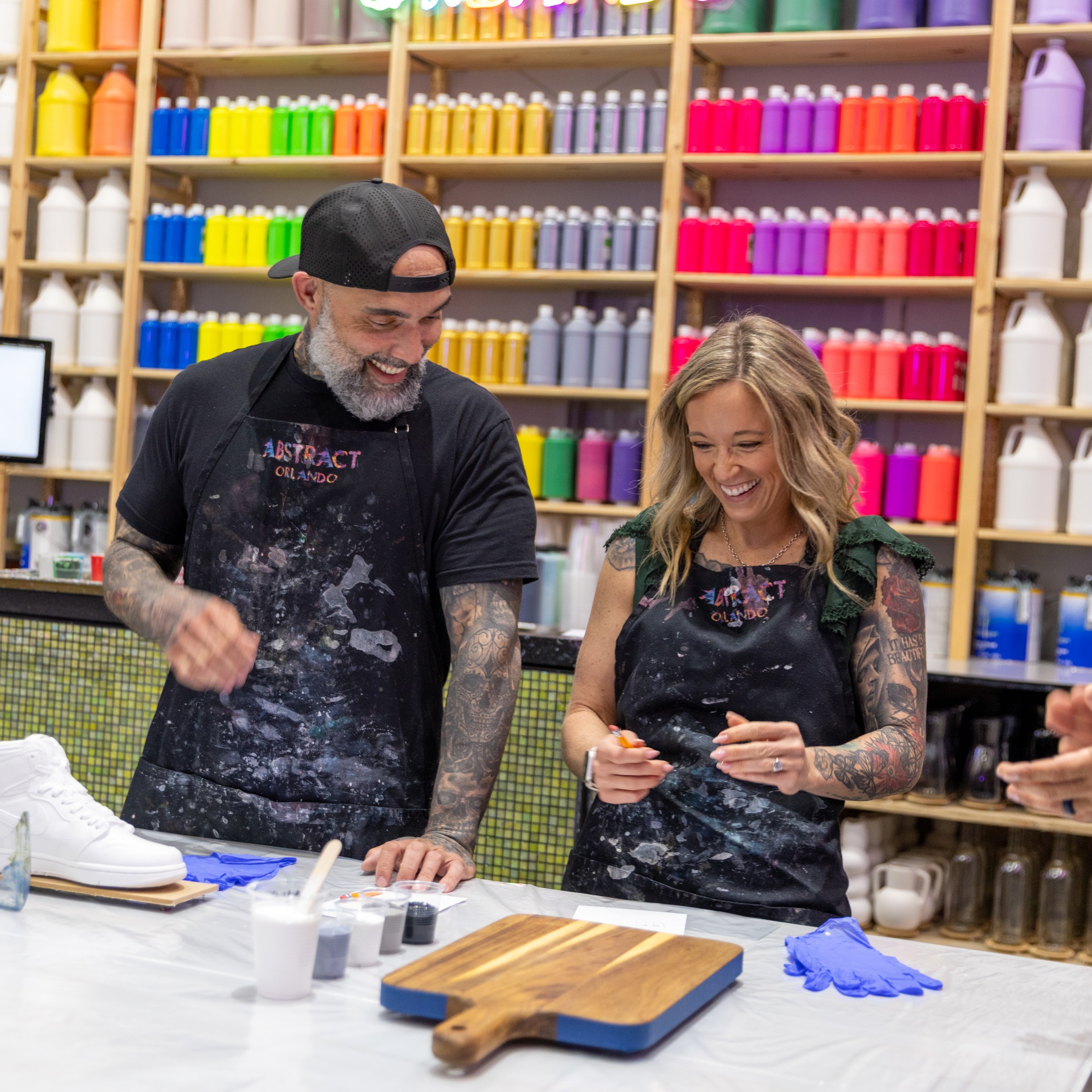 Two people in aprons painting in a colorful art studio with paint bottles on shelves.