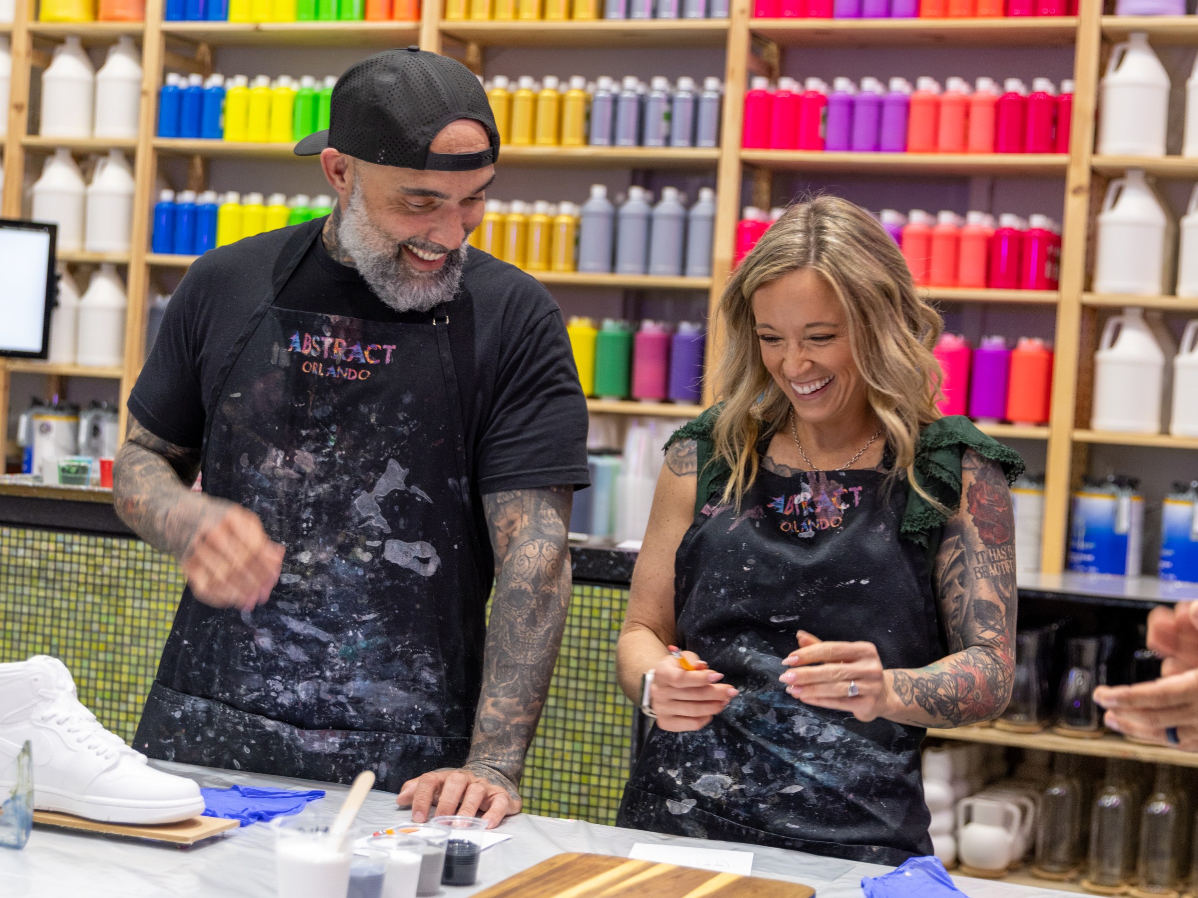 Two people in aprons painting in a colorful art studio with paint bottles on shelves.