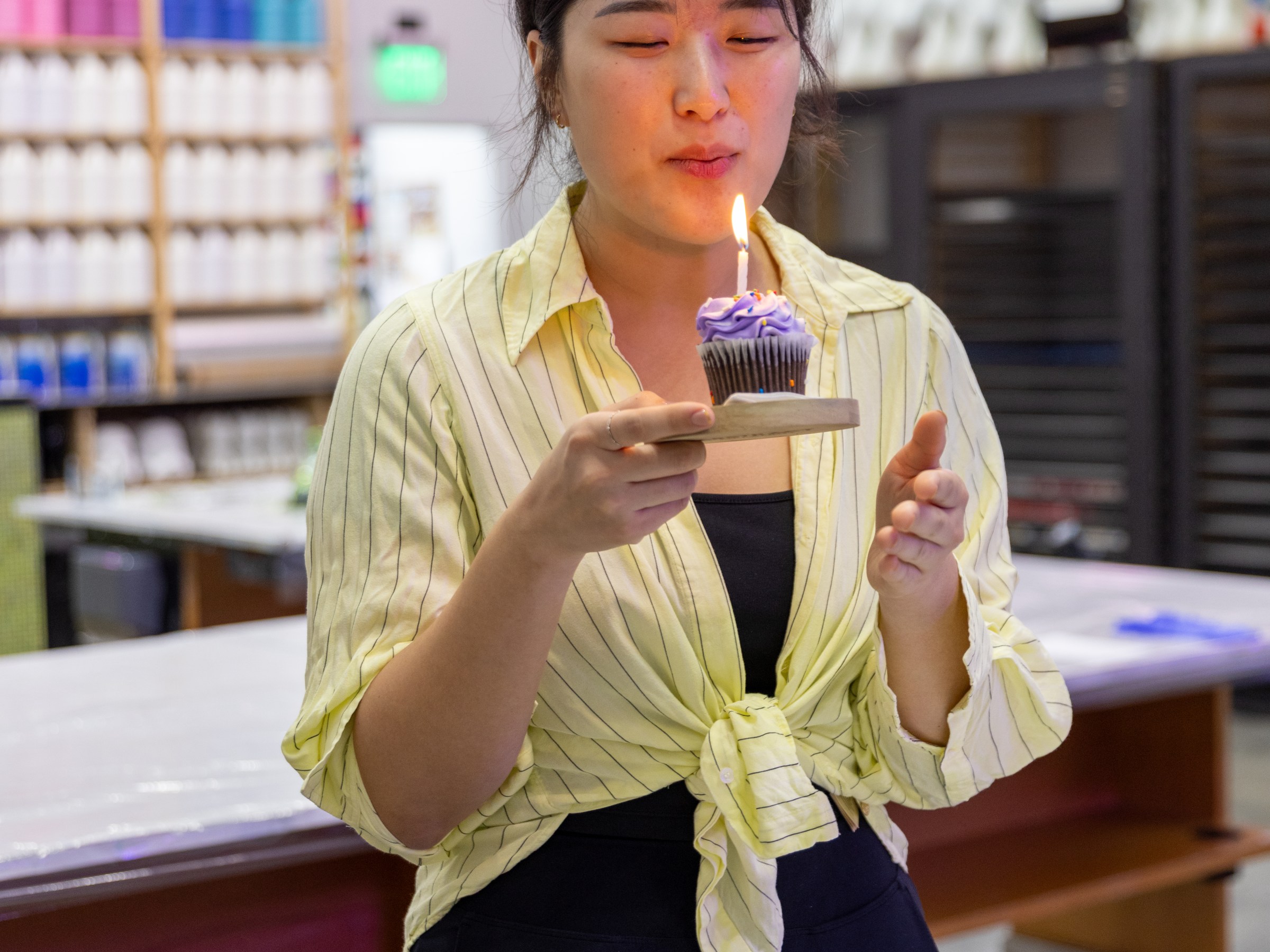 Person in a striped shirt holding a cupcake with a lit candle, blurred colorful background.
