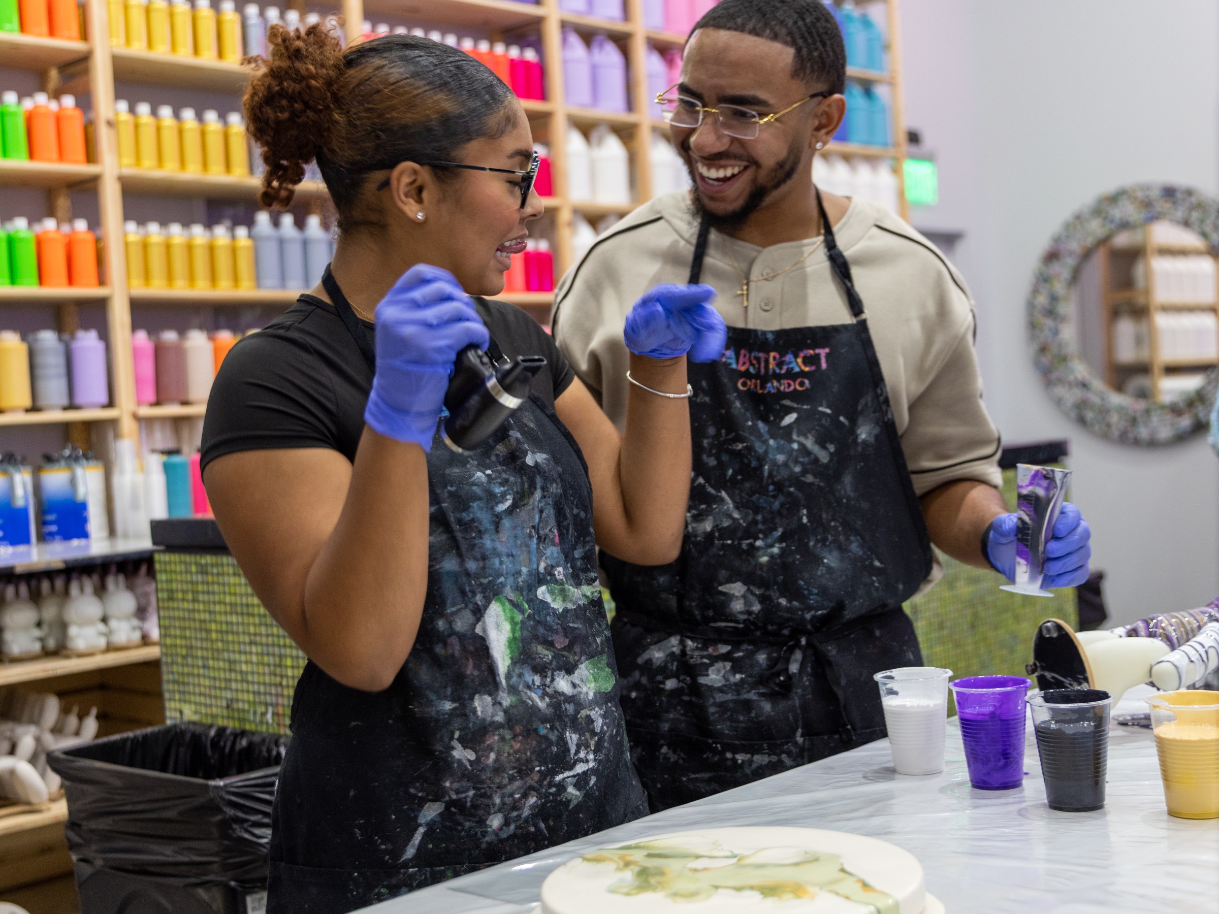Two people painting with colorful paints in an art studio, smiling and wearing aprons.