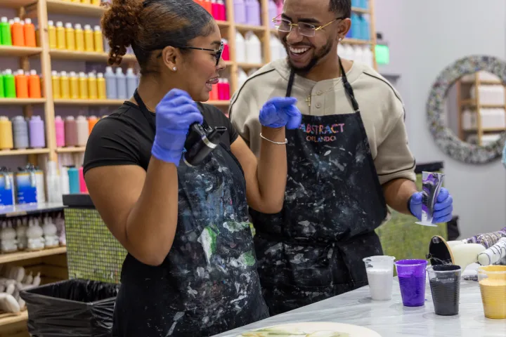 Two people painting with colorful paints in an art studio, smiling and wearing aprons.