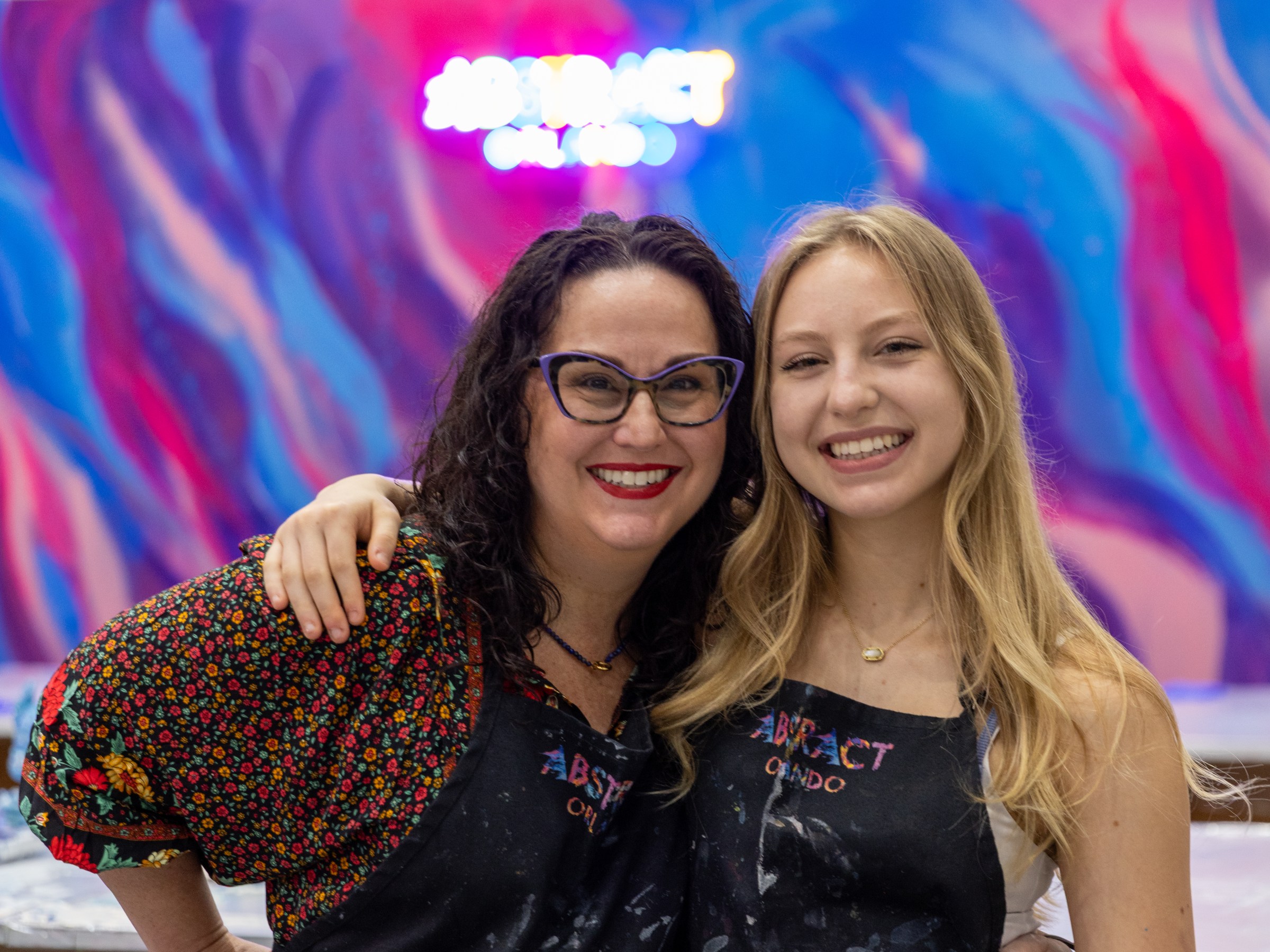 Two smiling women in aprons posing in front of a colorful backdrop with a decorated tree.