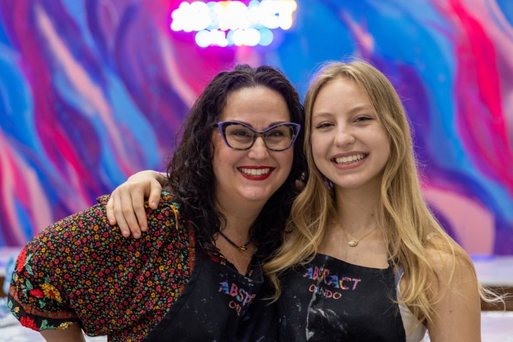 Two smiling women in aprons posing in front of a colorful backdrop with a decorated tree.