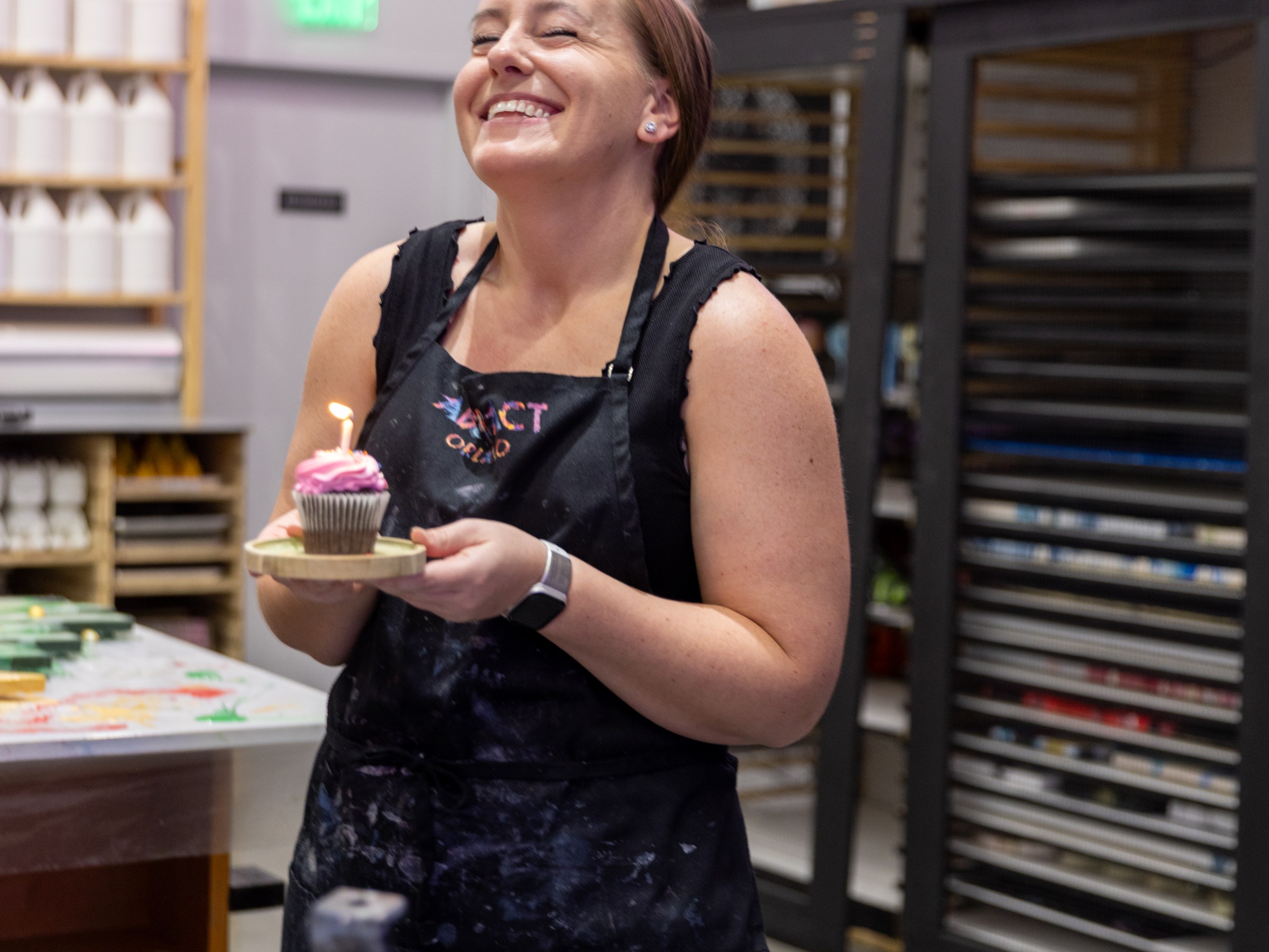 Woman in apron smiling, holding a cupcake with a lit candle in an art studio.