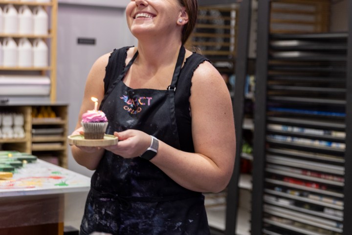 Woman in apron smiling, holding a cupcake with a lit candle in an art studio.