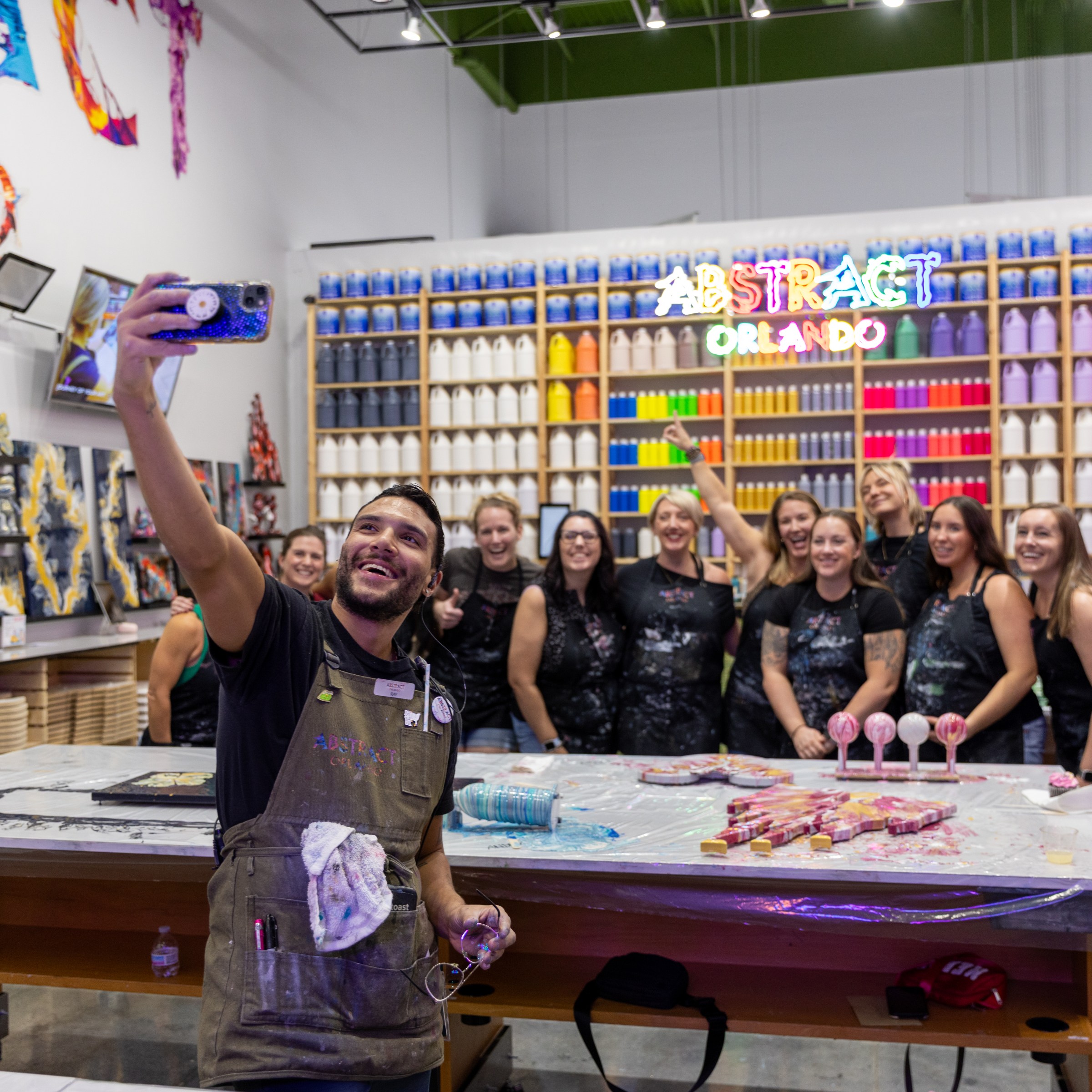 Group photo at art studio with colorful paint shelves and neon sign reading 'Abstract Orlando.'