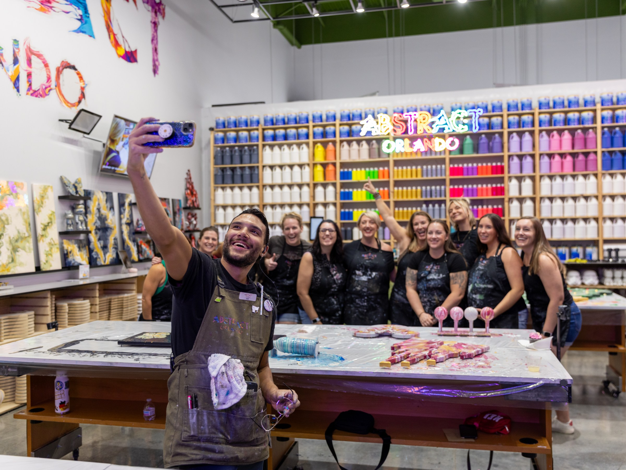 Group photo at art studio with colorful paint shelves and neon sign reading 'Abstract Orlando.'