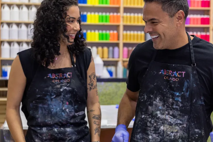 Two people smiling and painting in an art studio with colorful bottles and a neon sign in the background.