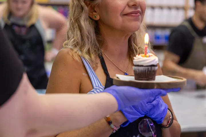 Smiling woman holds a cupcake with lit candle, surrounded by colorful paint bottles.