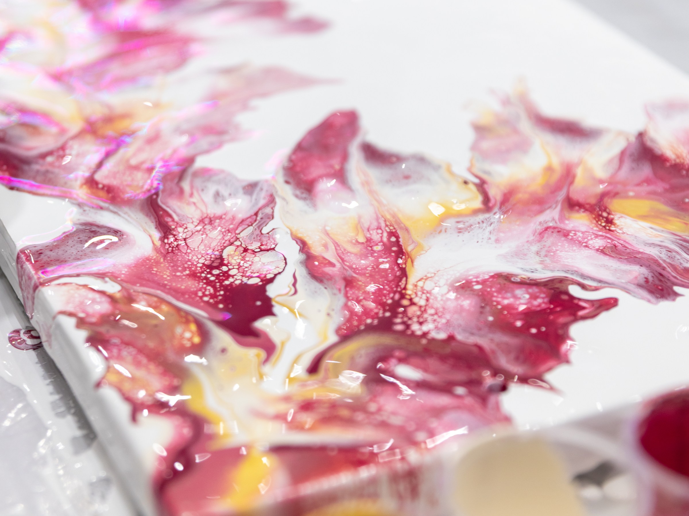 Close-up of abstract pink and yellow paint on a canvas with paint cups in foreground.