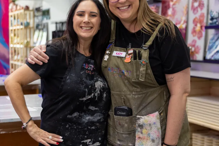 Two women in aprons smiling in an art studio with paintings displayed.
