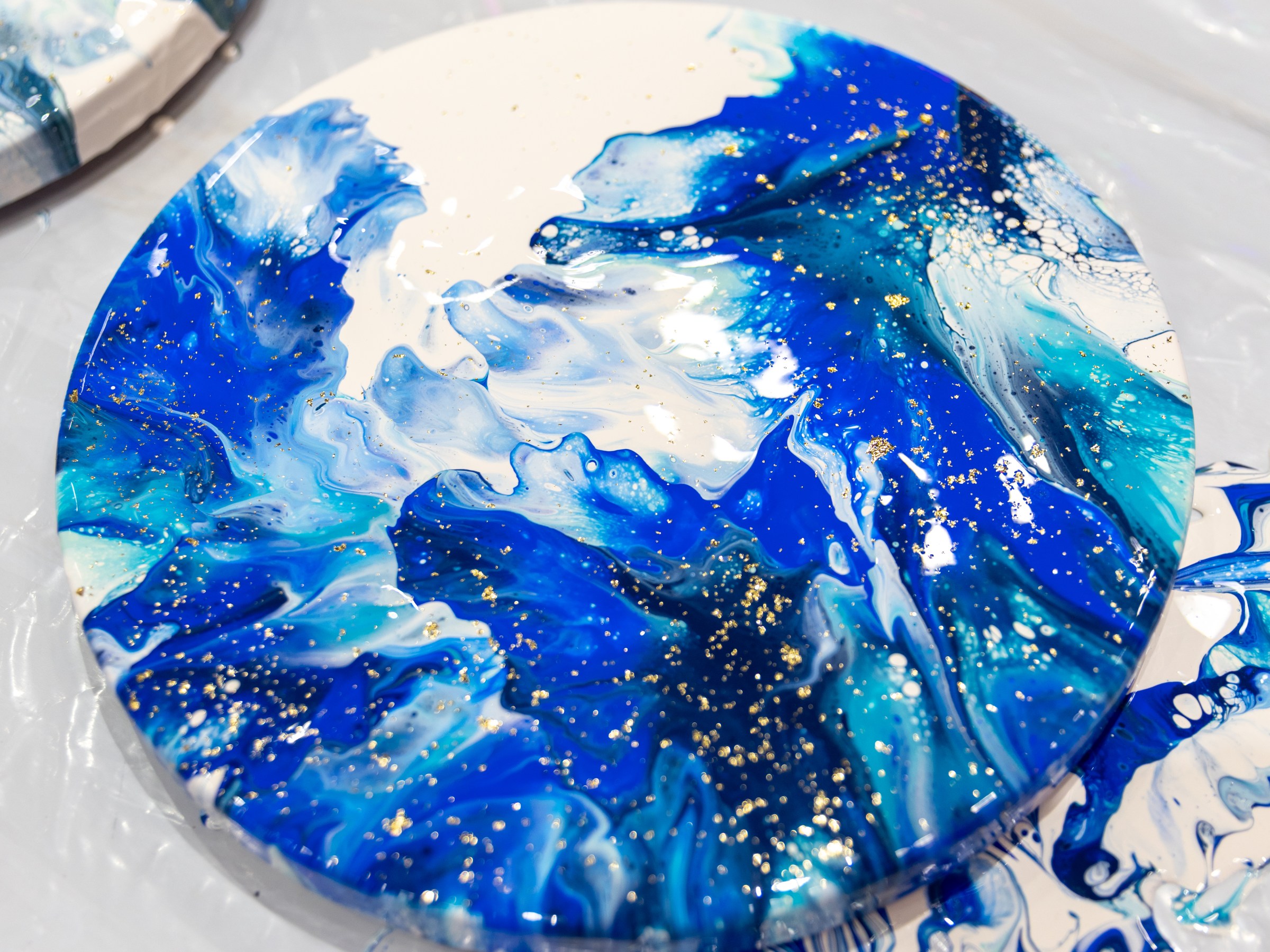 Round canvas with blue, white, and gold abstract fluid art on a plastic-covered surface.