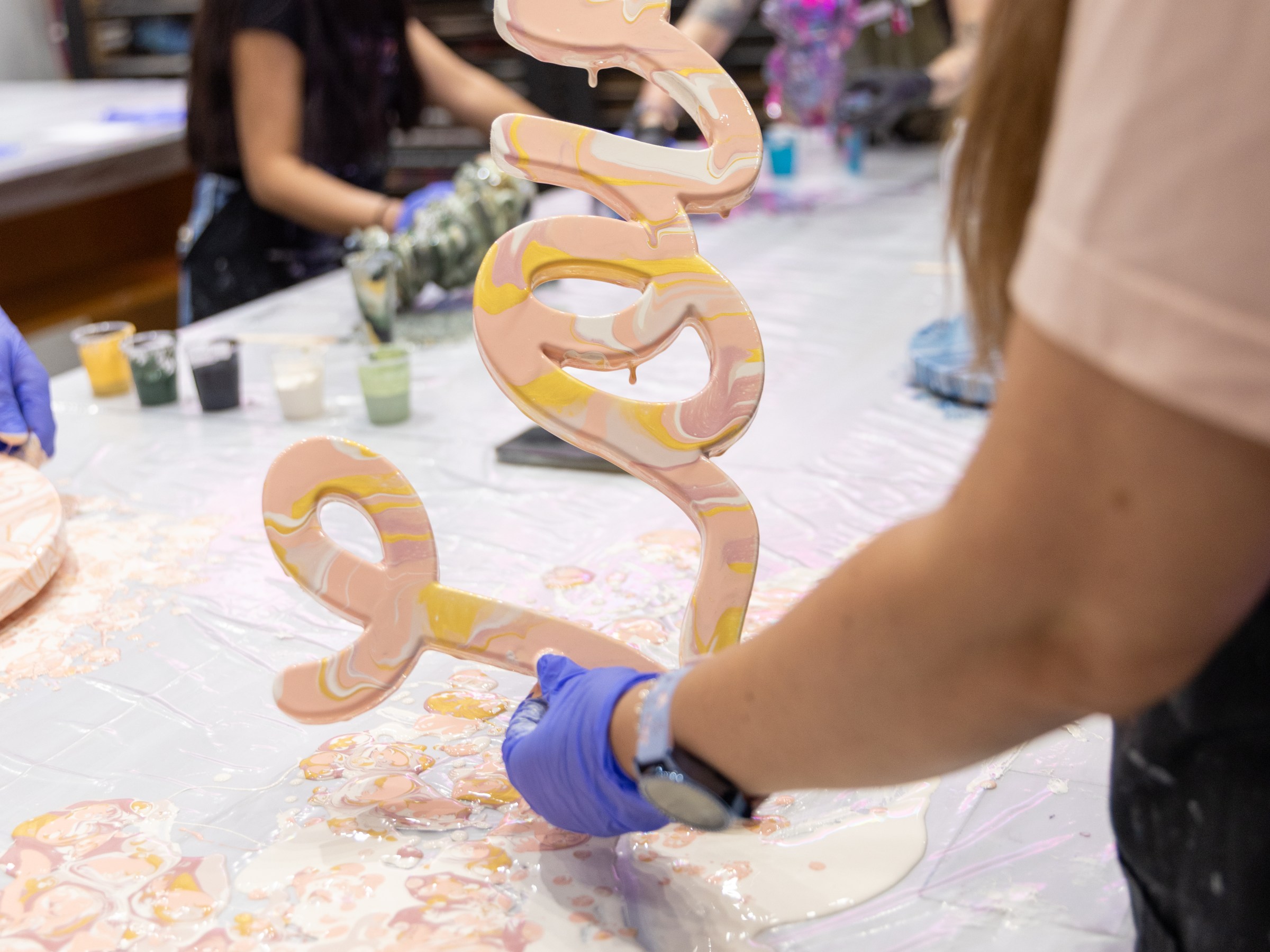 Person in gloves holding a wavy, colorful art piece over a messy table.