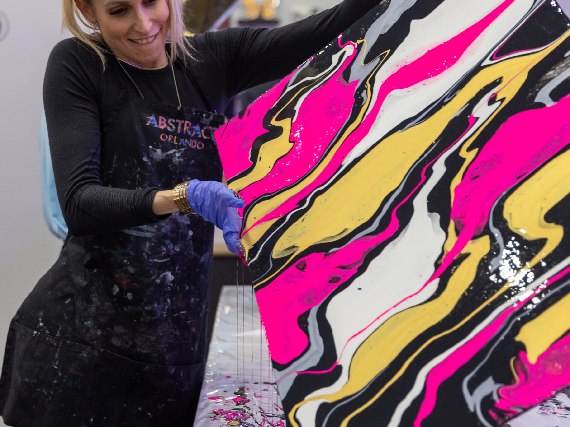 Person creating abstract art with pink, black, yellow, and white paint on a canvas.