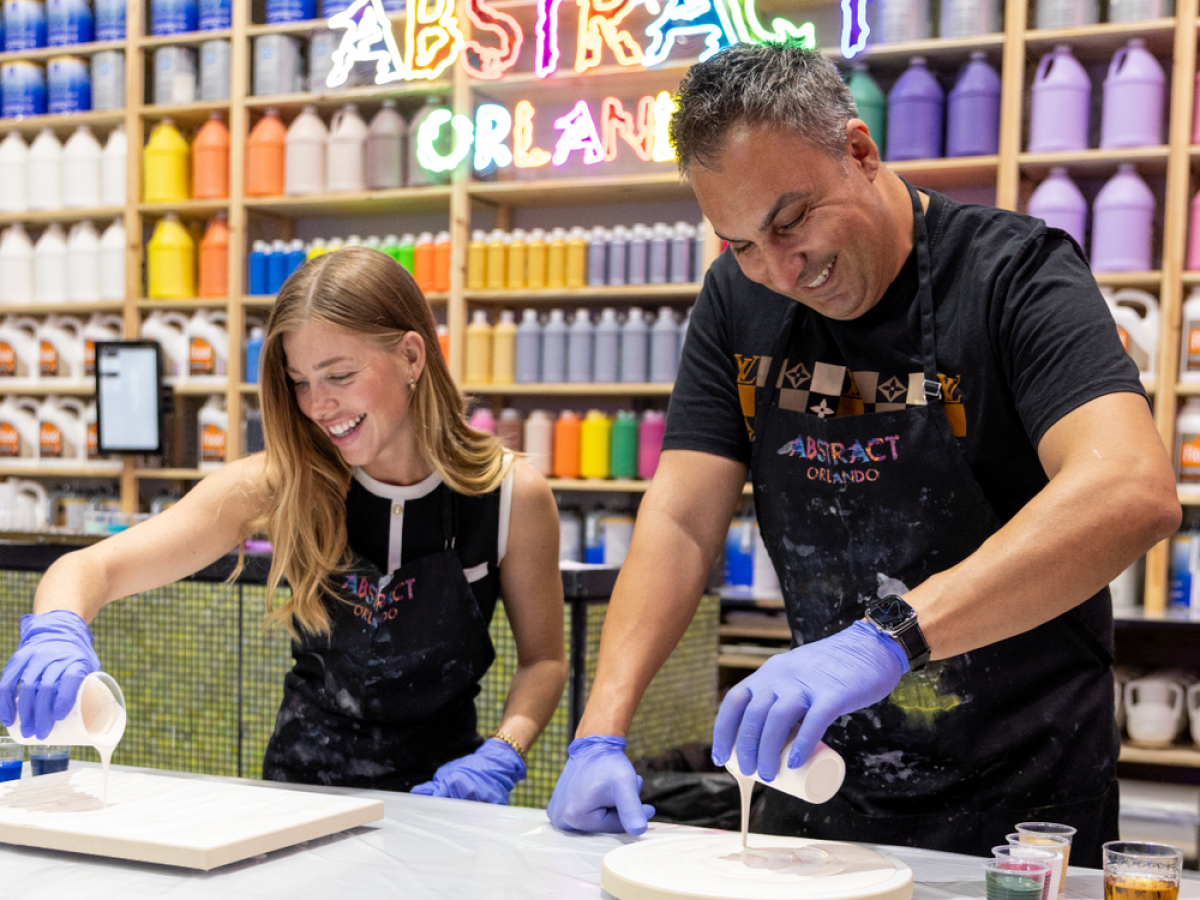 Two people pouring paint on canvases, wearing gloves, with paint bottles in the background.