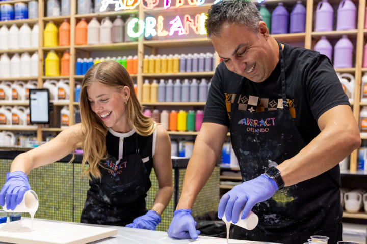 Two people pouring paint on canvases, wearing gloves, with paint bottles in the background.
