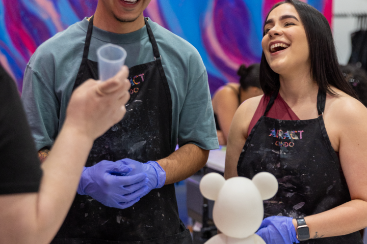 Two people smiling, wearing aprons, with paint cups and a bear-shaped figurine on a table.