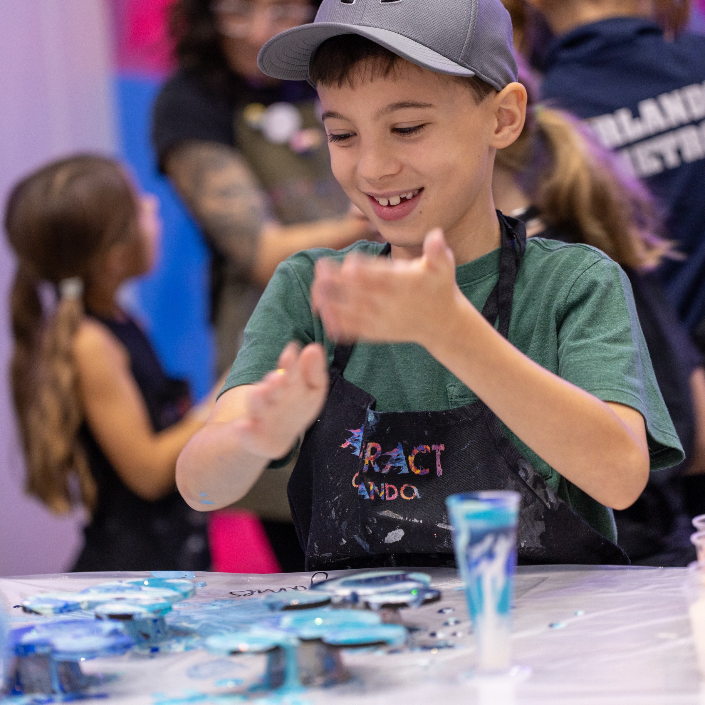 Boy in cap and apron smiles while painting at table with blue colors.