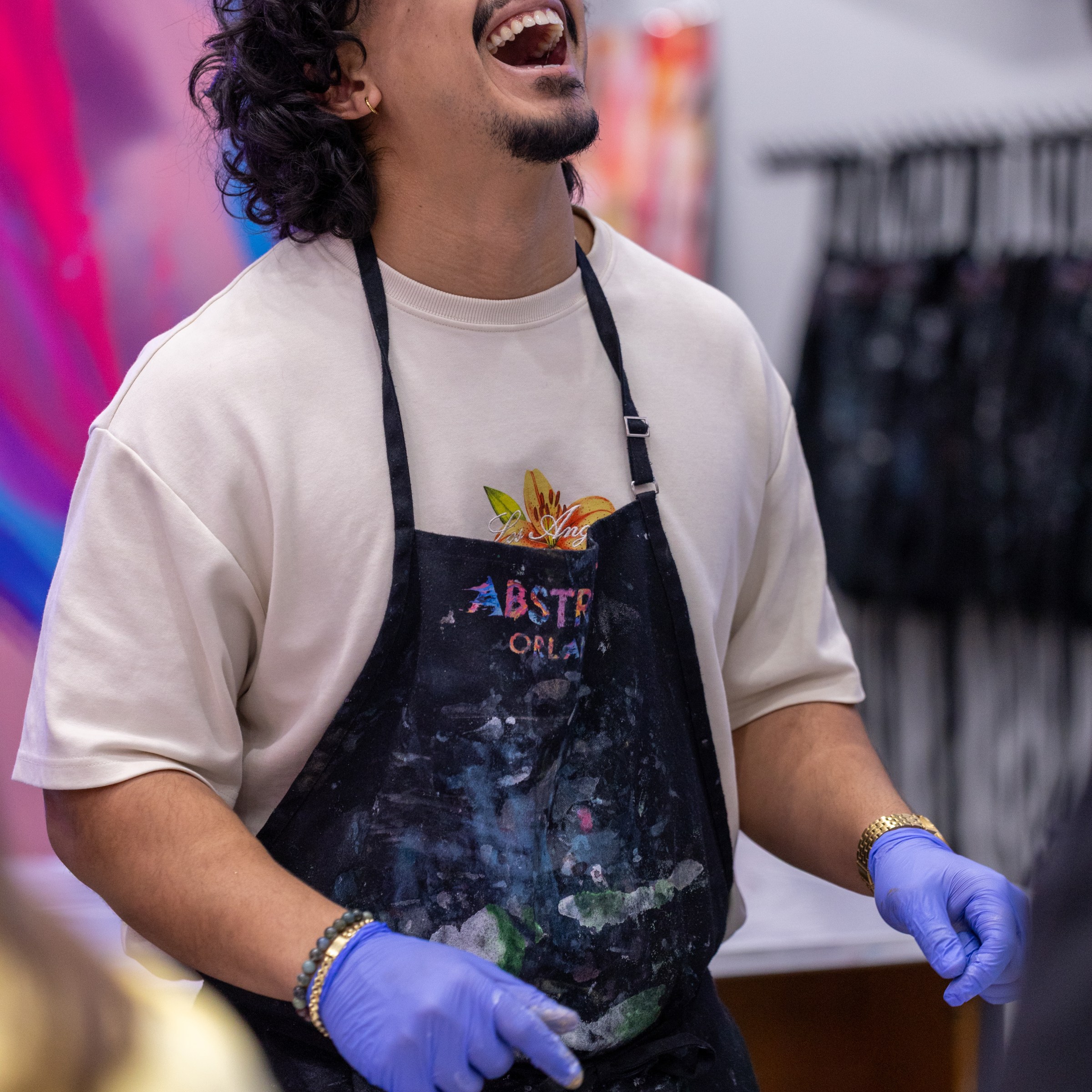 Artist laughing with eyes closed, wearing gloves and paint-stained apron near colorful artwork.
