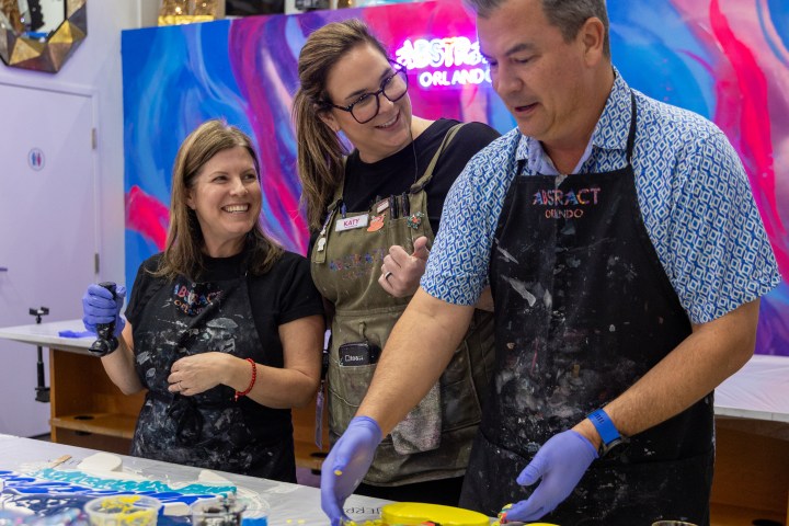 Three people in aprons and gloves painting a colorful abstract art piece on a table.
