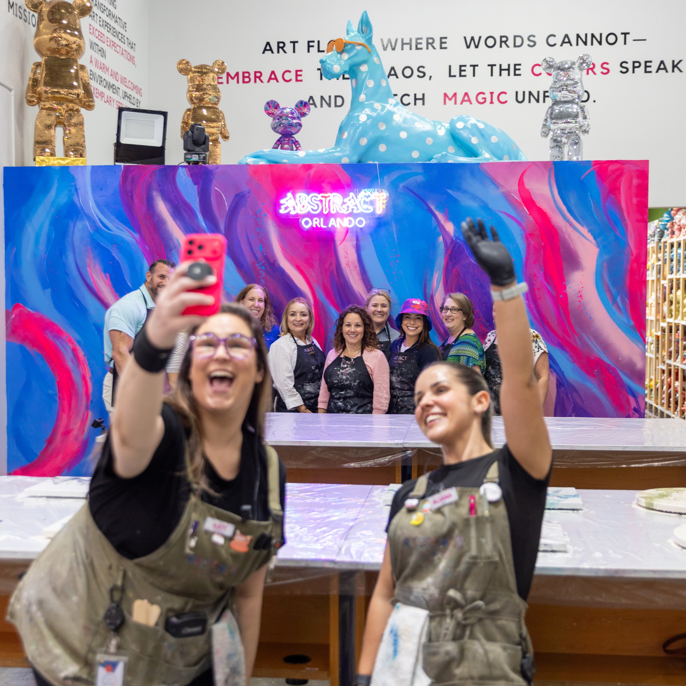 Two women taking a selfie with a colorful art backdrop and a group of people behind them.