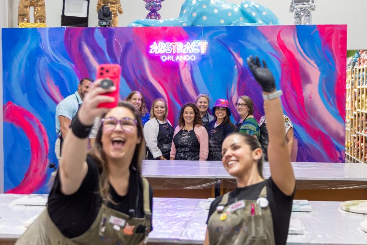 Two women taking a selfie with a colorful art backdrop and a group of people behind them.
