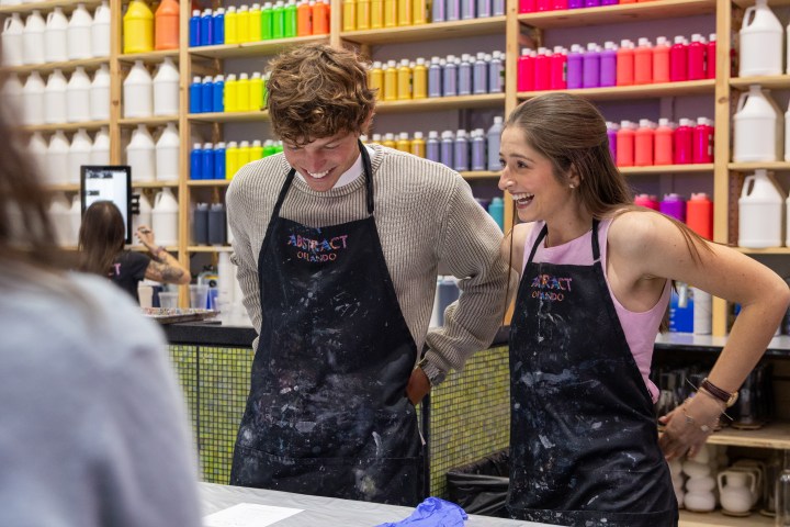 Two people in aprons smile at a paint studio with colorful shelves and neon 'Abstract Orlando' sign.