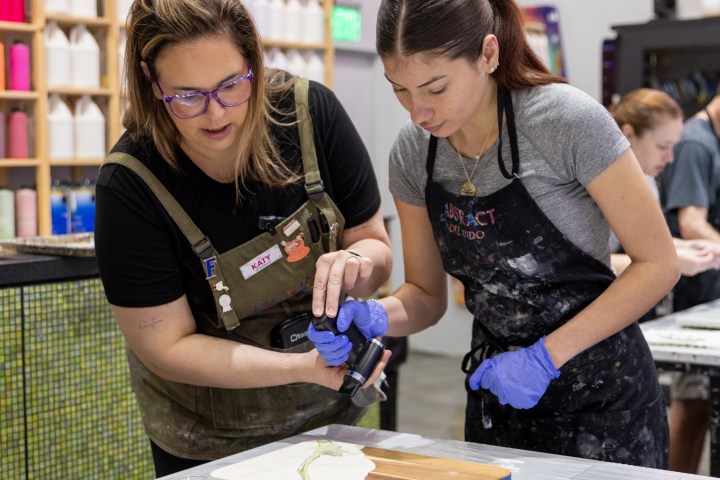 Two women in aprons and gloves work on a painting in a colorful art studio.