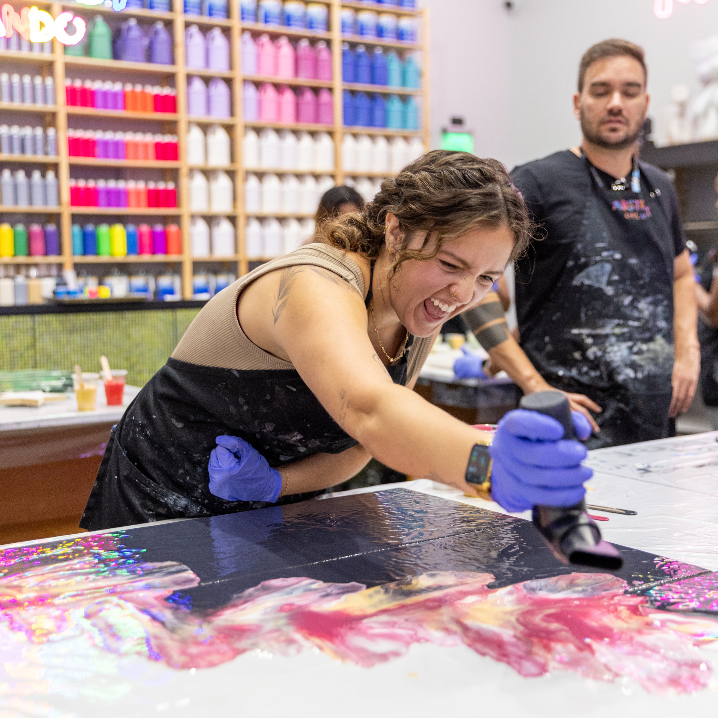 Woman joyfully painting with a heat gun, colorful bottles in background.
