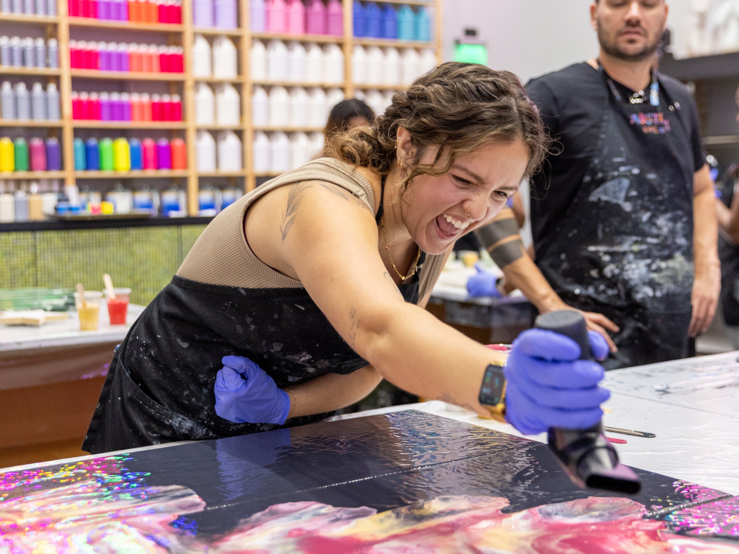 Woman joyfully painting with a heat gun, colorful bottles in background.