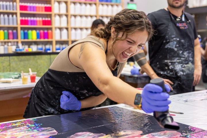 Woman joyfully painting with a heat gun, colorful bottles in background.
