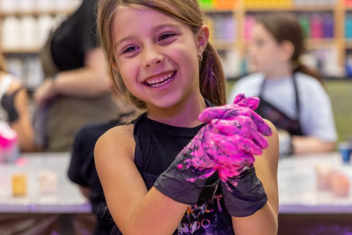 Smiling girl in an art class with pink paint on gloves, background of colorful paint shelves.