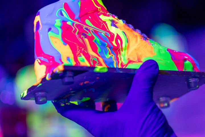 Gloved hands hold a sneaker covered in vibrant neon paint under UV light.