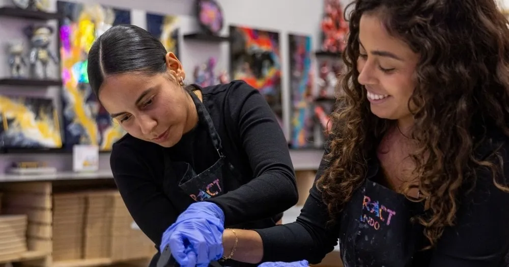 0c4ca870e73a807a0992686c70392f8756264c6b Two women in black shirts painting together, smiling, with art on walls behind.
